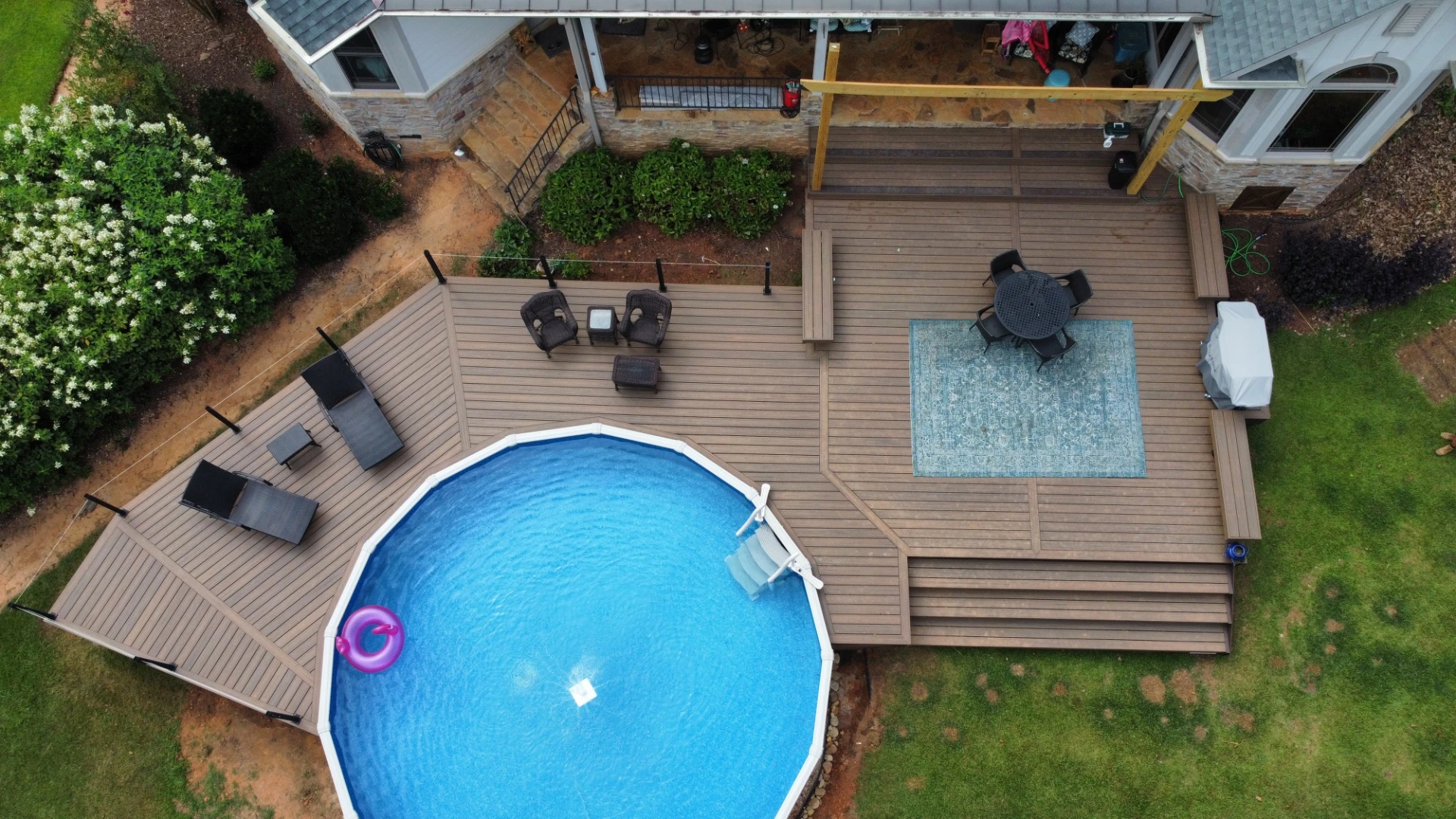Aerial view of a backyard circle shape pool surrounded by a large wooden deck and patio seating